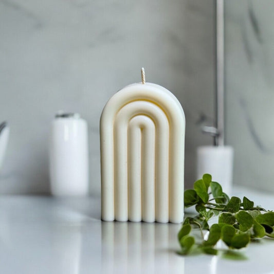 Bougie Arc-en-ciel blanche en forme d’arche nervurée, posée sur une table grise près d’une plante verte