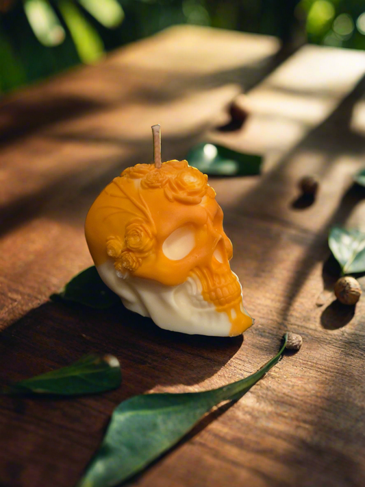 Bougie tête de mort orange et blanche ornée de roses, vue de trois quarts sur une table en bois avec des feuilles.