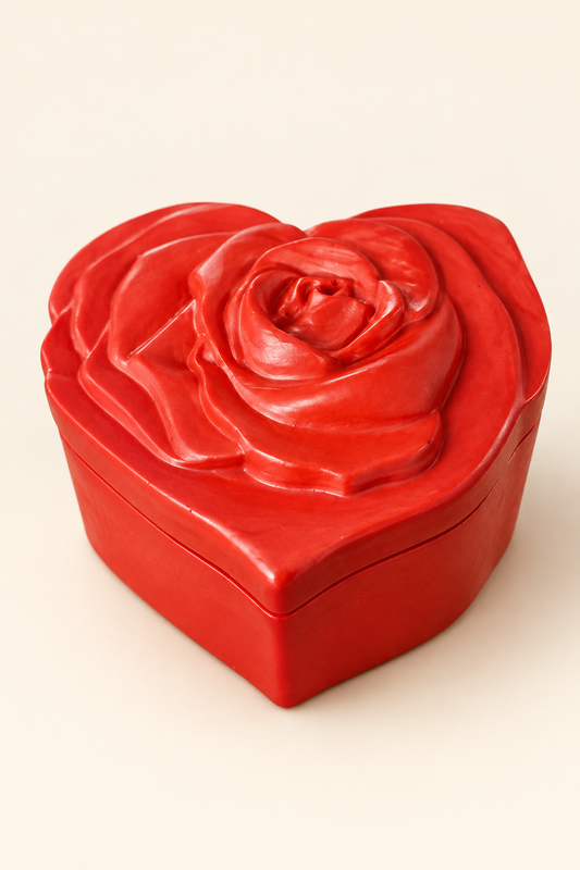 Boîte rouge en forme de cœur, fermée avec un couvercle orné d’une rose en relief, photographiée sur fond ivoire.