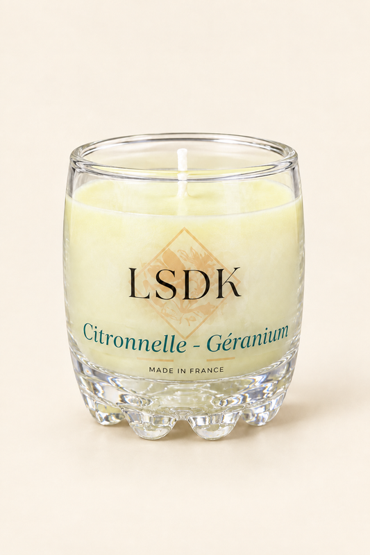 Bougie LSDK Citronnelle et Géranium non allumée, dans un verre transparent, photographiée sur fond beige.