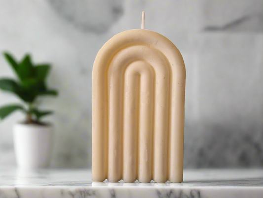 Bougie Arc-en-ciel beige en forme d’arche nervurée, dans un décor clair avec une plante. 