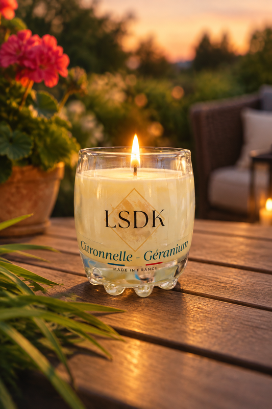 Bougie LSDK Citronnelle et Géranium allumée dans un verre, sur une table en bois en terrasse au coucher du soleil.
