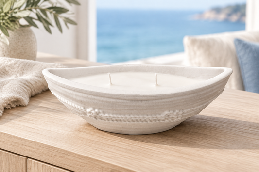 Bougie blanche en forme de barque avec deux mèches et un relief de corde, sur un meuble en bois devant une vue sur la mer.
