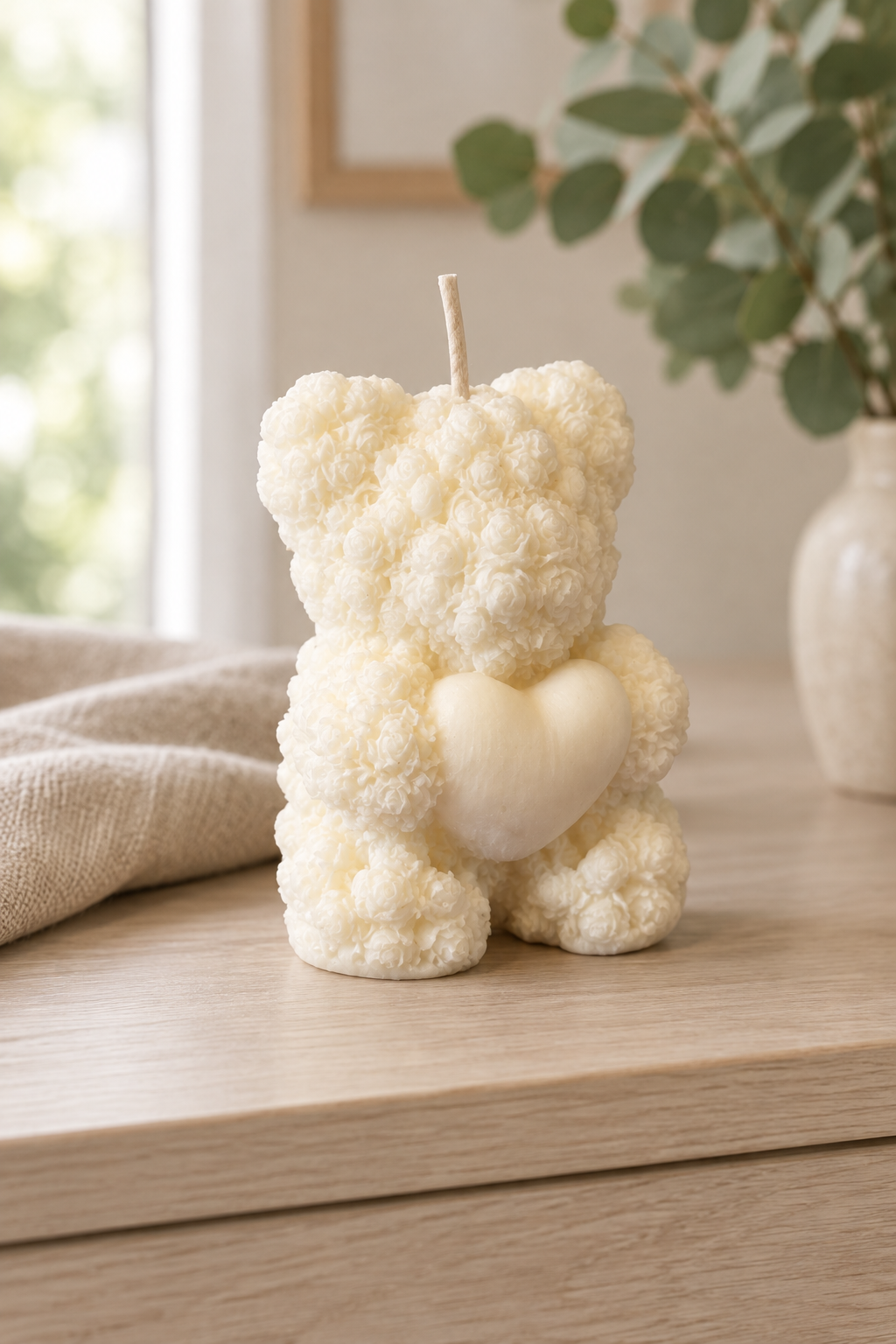 Bougie ivoire en forme d’ourson au relief de petites roses, tenant un cœur, avec une mèche, sur un meuble en bois clair.
