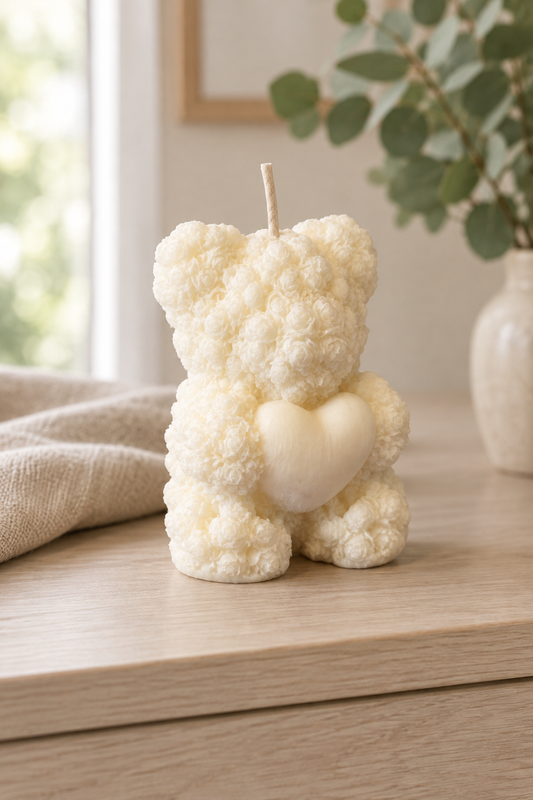 Bougie ivoire en forme d’ourson au relief de petites roses, tenant un cœur, avec une mèche, sur un meuble en bois clair.
