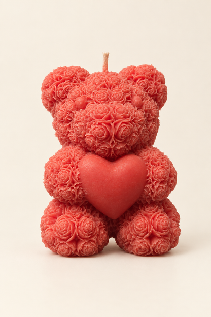 Bougie rouge en forme d’ourson au relief de petites roses, tenant un cœur, avec une mèche, photographiée sur fond ivoire.
