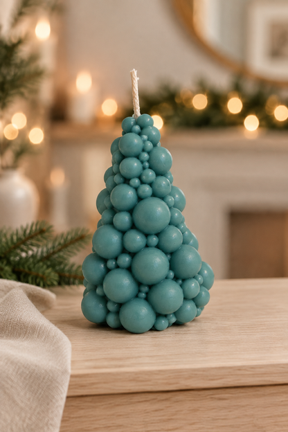 Bougie vert bleuté en forme de sapin composé de boules, sur un meuble en bois avec des branches de sapin et des lumières floues.
