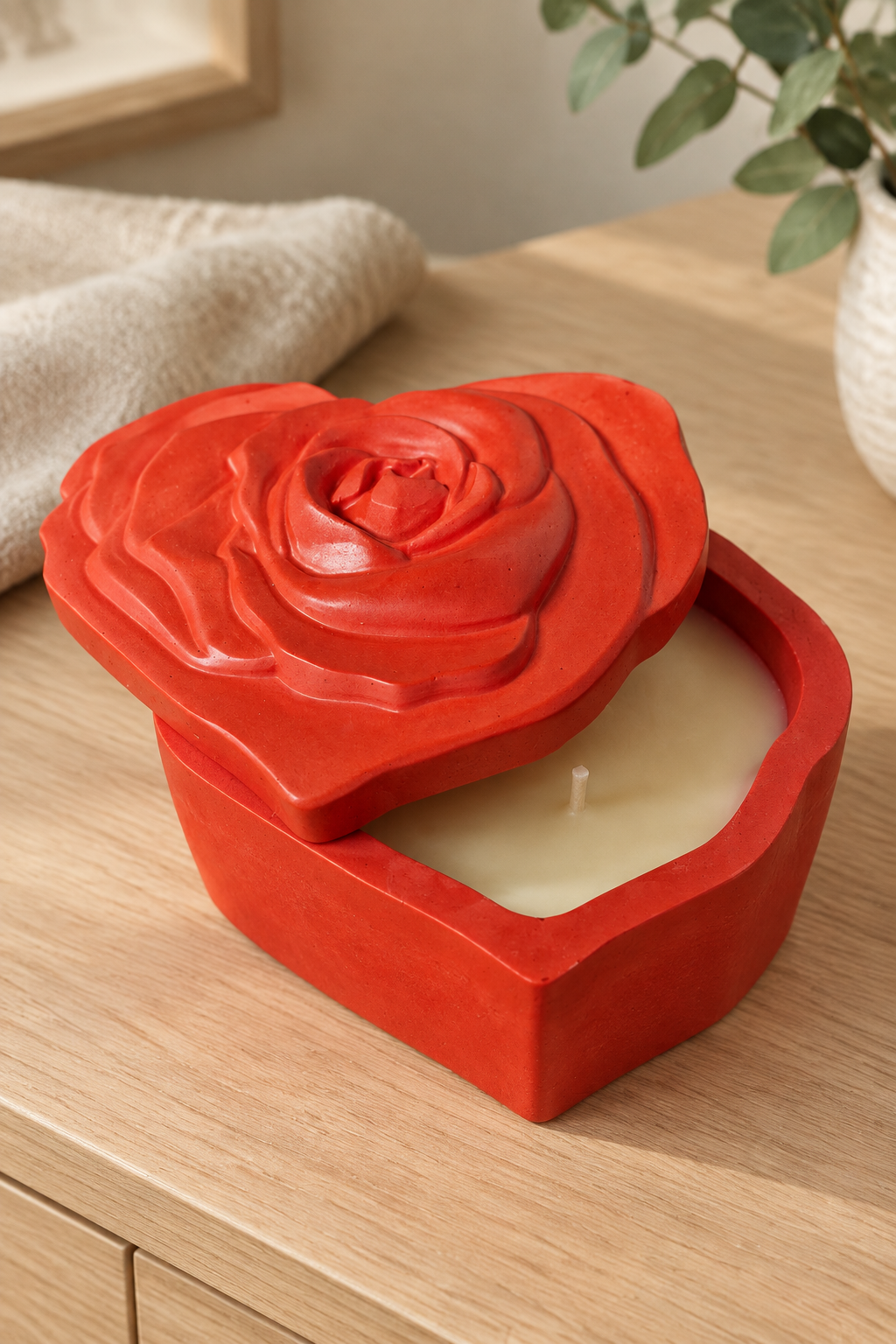 Bougie rouge en forme de cœur, couvercle à rose en relief entrouvert laissant voir la cire et la mèche, sur un meuble en bois.
