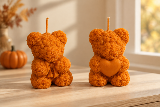 Deux bougies oursons orange au relief de roses, l’un avec un nœud et l’autre un cœur, sur un meuble en bois avec une citrouille.
